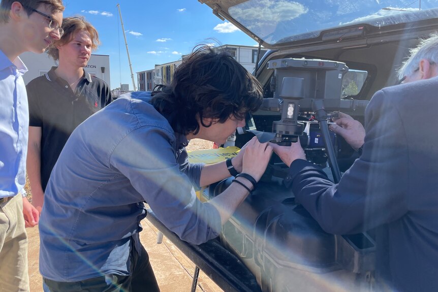 Three male univesity students look at a large black drone which sits in back of a ute