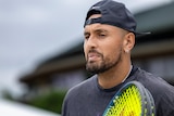 A pensive Nick Kyrgios looks down as he stands on a practice court with his racquet.
