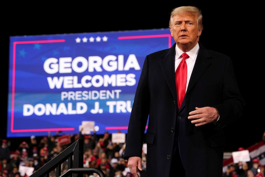 Donald Trump stands on stage in a suit and tie in front of a large screen that says 'Georgia welcomes President Donald J Trump'