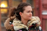 A woman with a large coat on looks on, away from the camera, as snow falls