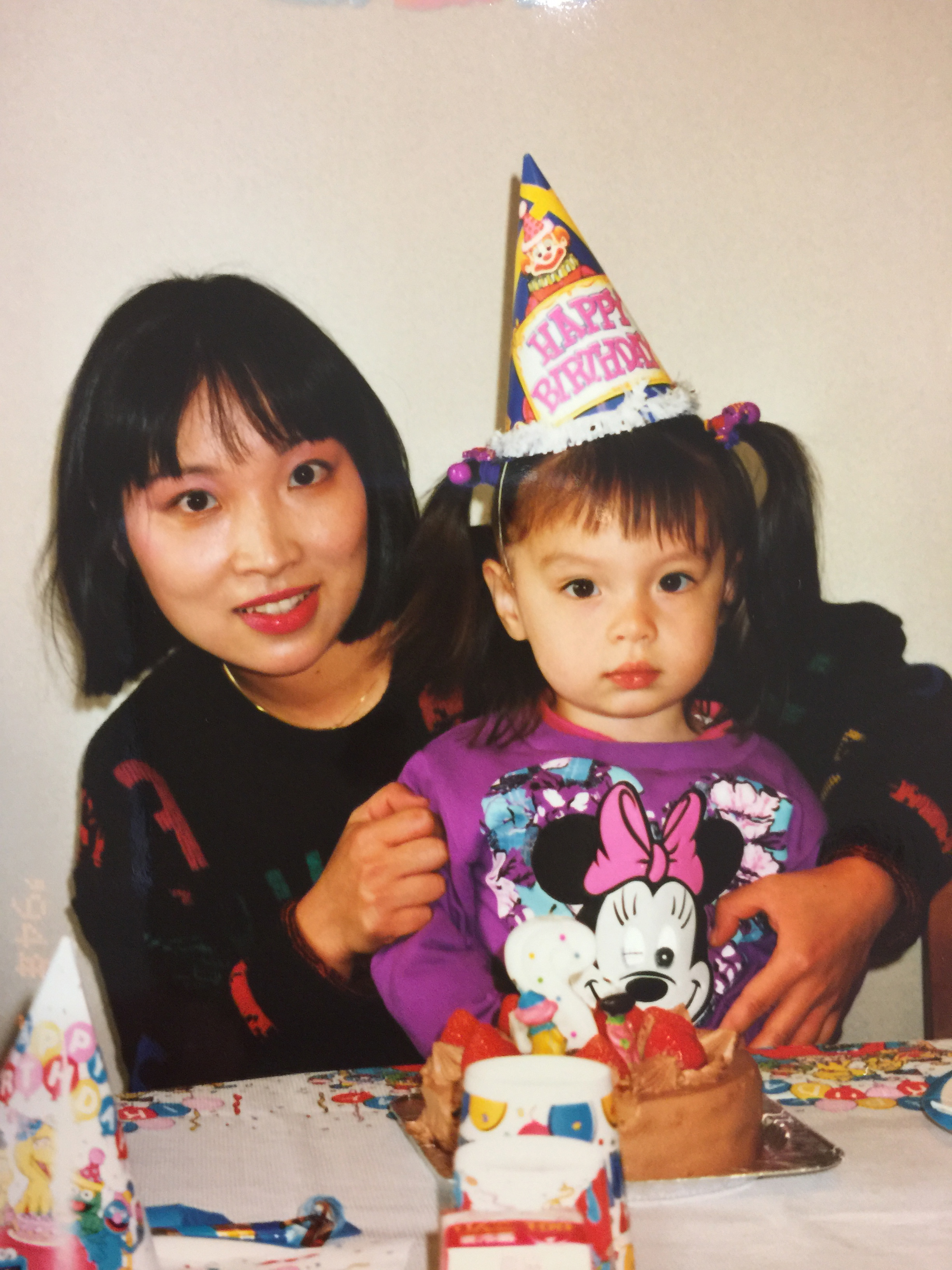 A Taiwanese woman with dark shoulder-length hair wears a jumper and holds a little girl in a birthday hat and Minnie Mouse top.