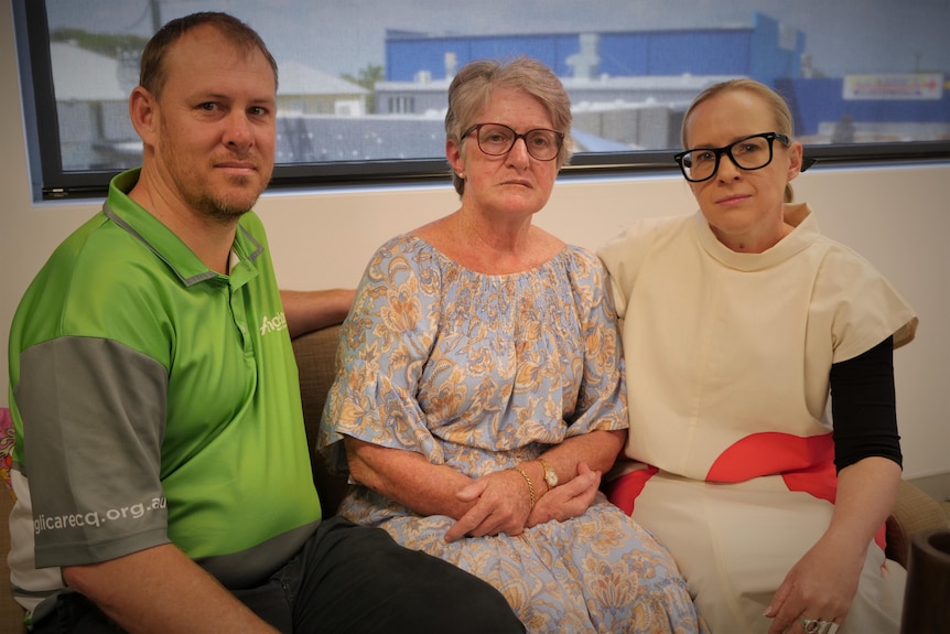 A man in a green shirt, next to two women with glasses, looking sombre at the camera.