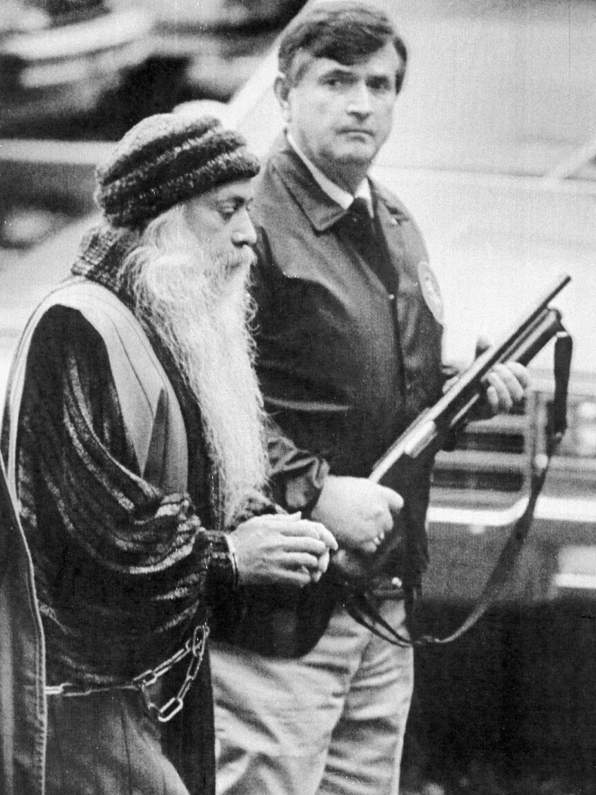 A man with a long white beard and handcuffs escorted by a middle-aged man holding a gun
