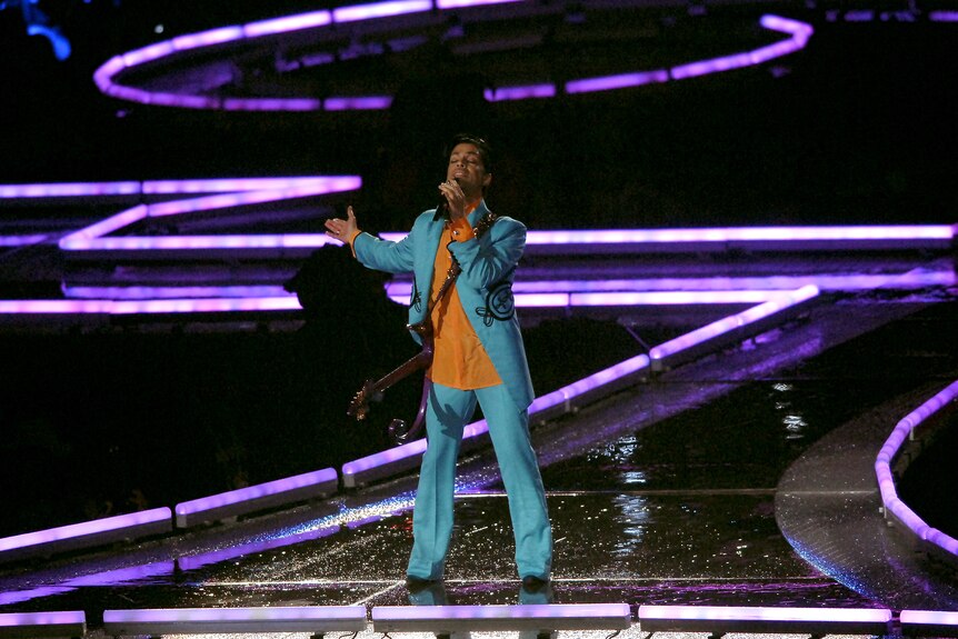 Prince on stage holding a microphone dressed in an aqua suit with orange shirt