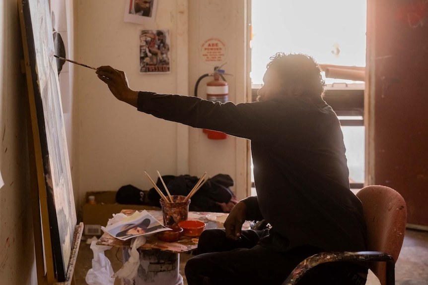 Vincent Namtjira holding a paintbrush up at the canvas, in the Iwantja Arts Studio