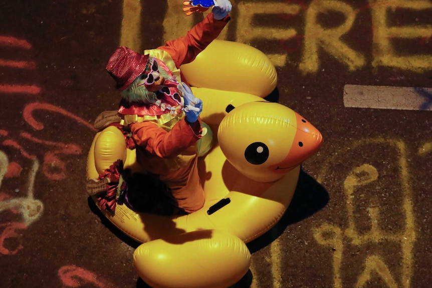 A clown kneels on massive inflatable duck and raises arms in the air.