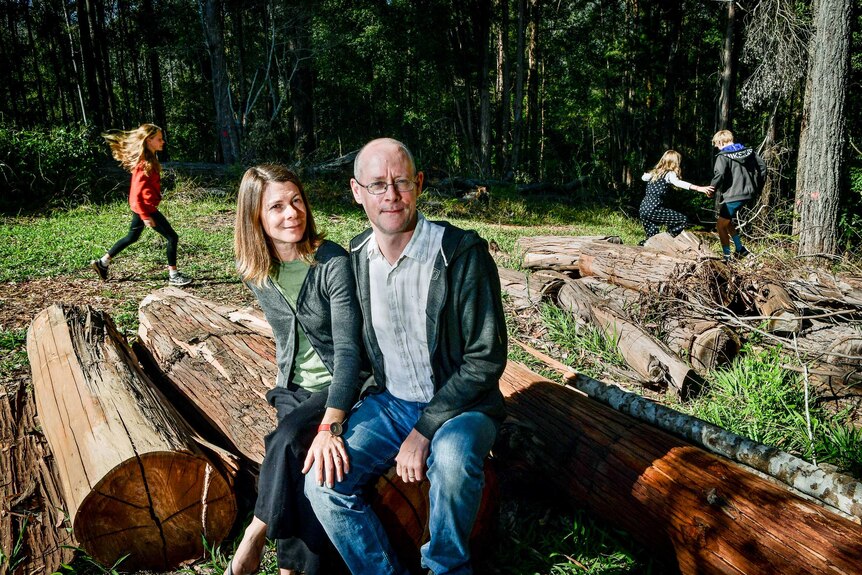 Mother and father sit on a log while three children run around in the background.