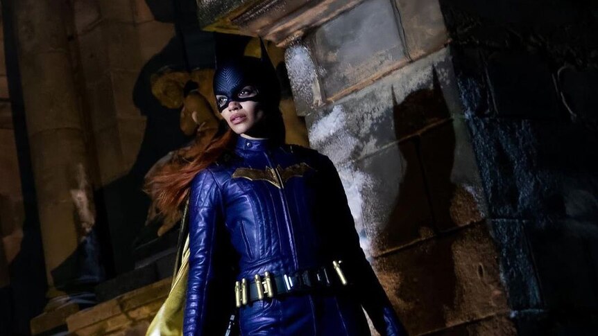 Batgirl stands solemnly by a stone pillar, casting a shadow onto it.
