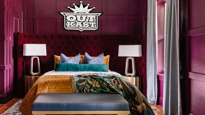 A bedroom with white and black OUTKAST logo above burgundy headboard, walls painted burgundy
