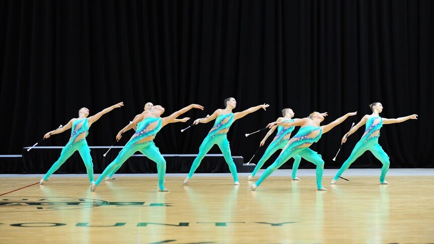 A group of women in green full body leotards lean to the left with their arms outstretched while holding batons.