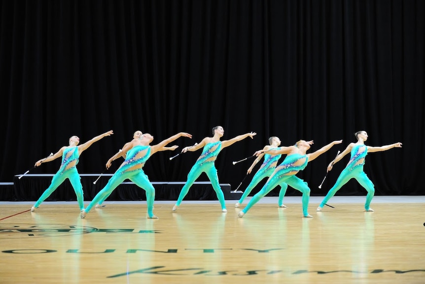 A group of women in green full body leotards lean to the left with their arms outstretched while holding batons.