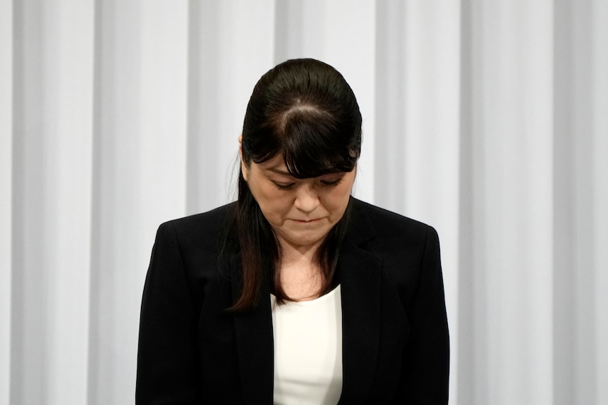 A woman wearing a blazer bows her head down with a solemn expression on her face
