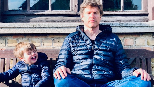 Steven Laureys sits on a wooden bench, in a cross-legged pose, eyes closed, meditating, while a child laughs next to him.