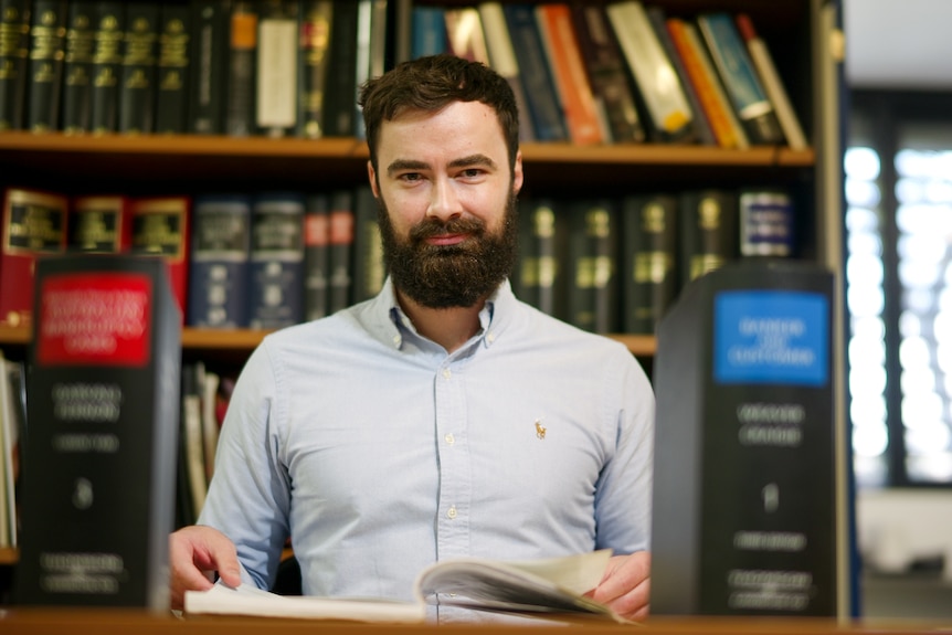 A man with dark hair and bushy beard sits in front of a wall of law books.