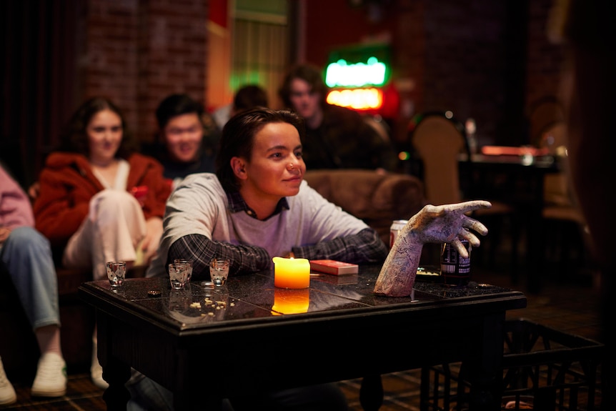 A non-binary millennial with short brown hair, sits leaning eagerly at a table with a creepy embalmed hand, surrounded by people
