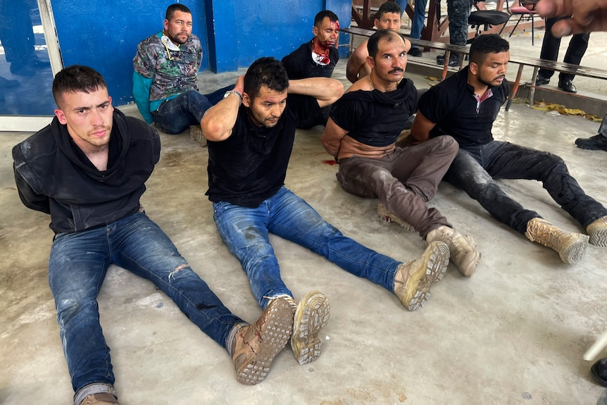 A group of men sit on the floor, some with their hands behind their backs, some with their hands behind their heads