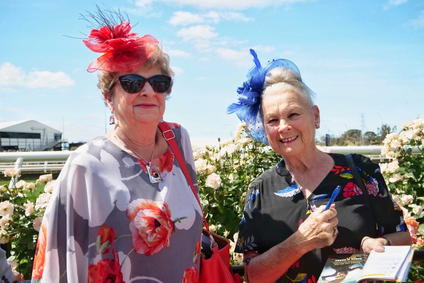 Two middle-aged women in colourful dresses and fascinators smile in front of the roses at Flemington Racecourse.