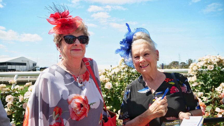 Two middle-aged women in colourful dresses and fascinators smile in front of the roses at Flemington Racecourse.