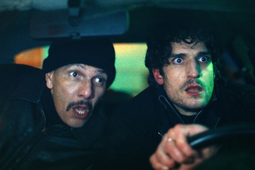 A middle-aged white man in a black skivvy and beanie looks stressed beside 30-something white man in black jacket in a car.