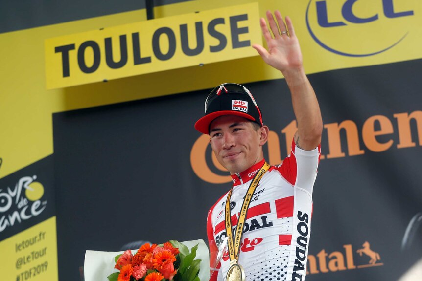 A cyclist with a medal around his neck, waves to the crowd from the podium at the Tour de France.