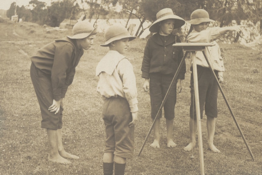 A black and white image showing four young boys wearing shirts, long pants and broad brimmed hats standing near a river bank.