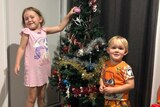 Two young kids stand in front of a Christmas tree