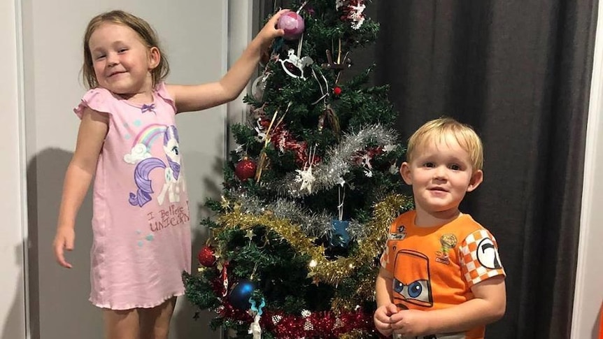 Two young kids stand in front of a Christmas tree