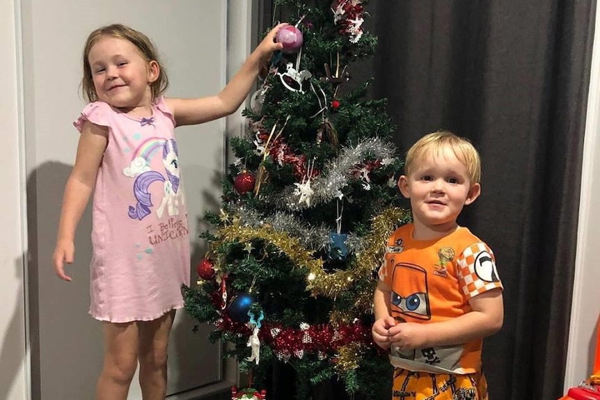 Two young kids stand in front of a Christmas tree