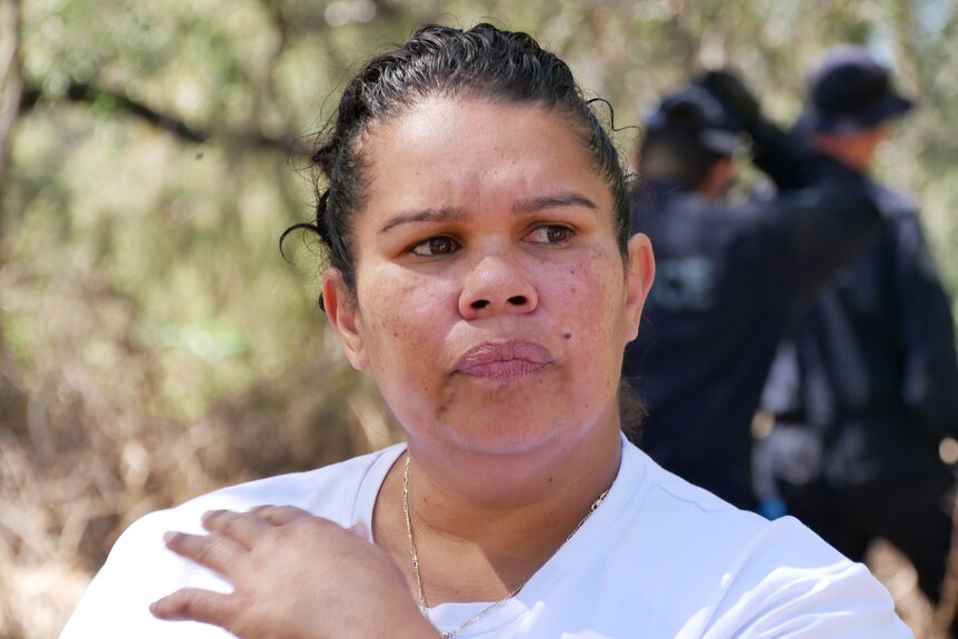 An Indigenous woman with a serious expression stands wearing a light top in the bush.