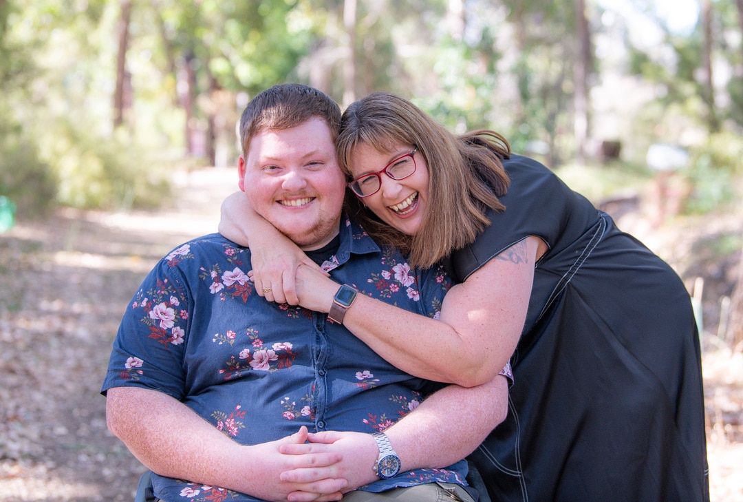Una madre y un sol se abrazan y sonríen a la cámara. Están afuera bajo el sol.