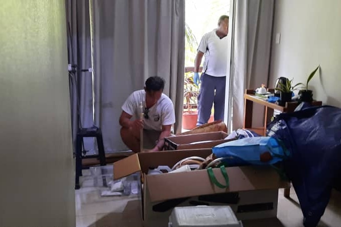 Australian man Jamie Pang kneeling on the ground, handling items in a hotel room. There is a man on the balcony behind him.