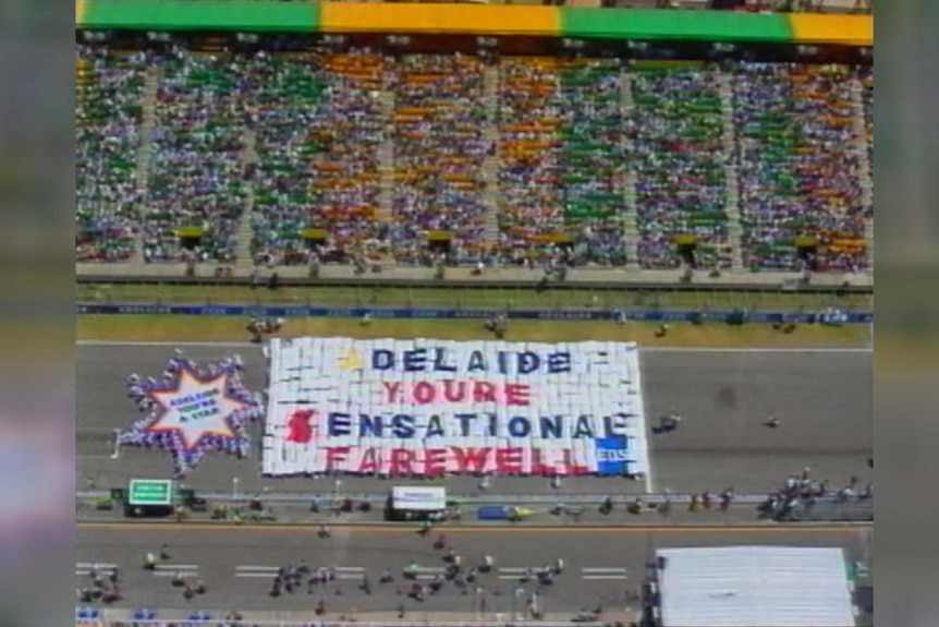 Archival vision of a race track with an enormous banner in front of the grandstand saying 'Adelaide You're Sensational Farewell'
