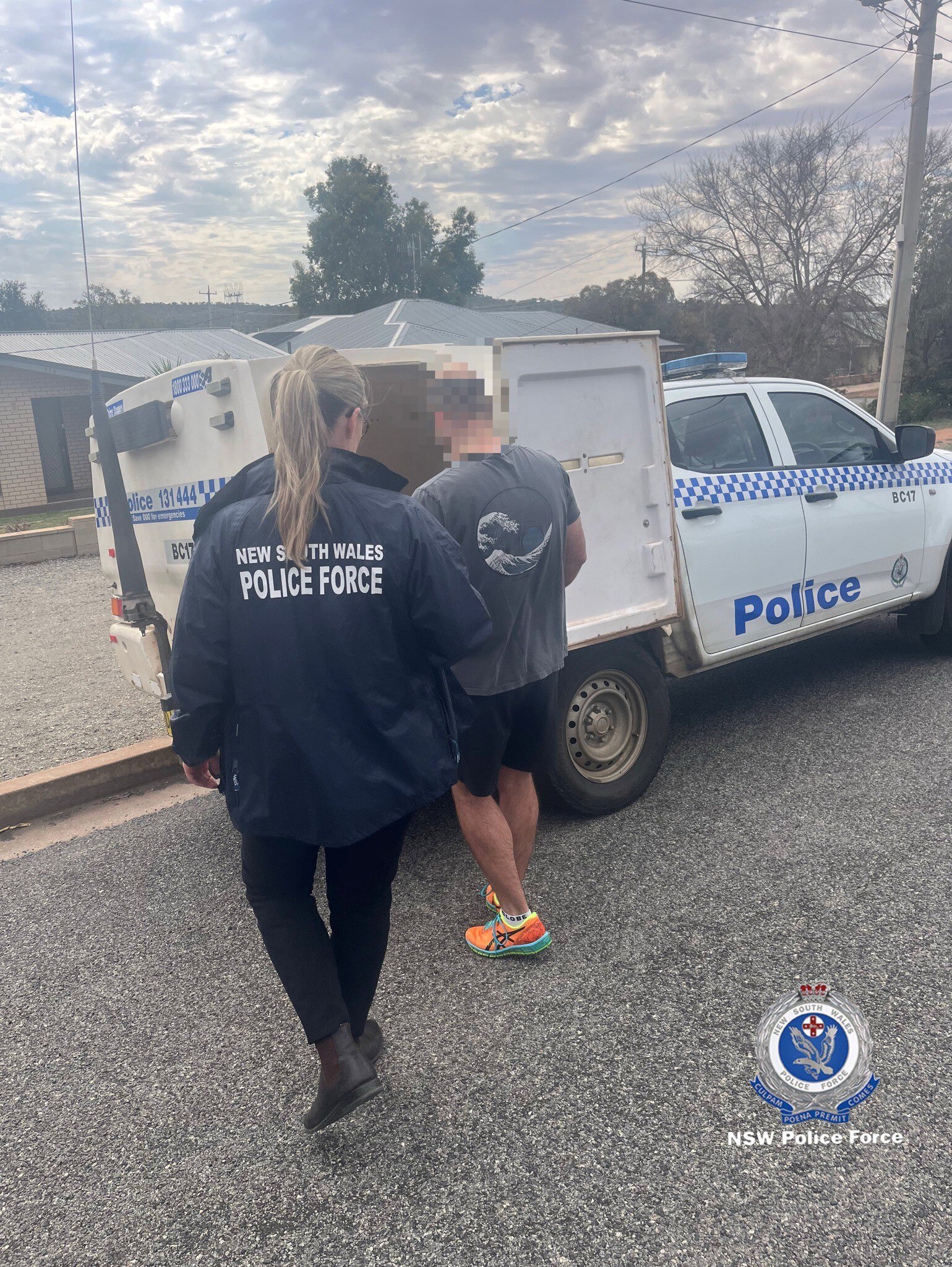A man wearing a t-shirt and shorts with his face blurred being led into a police paddy wagon by a female officer.