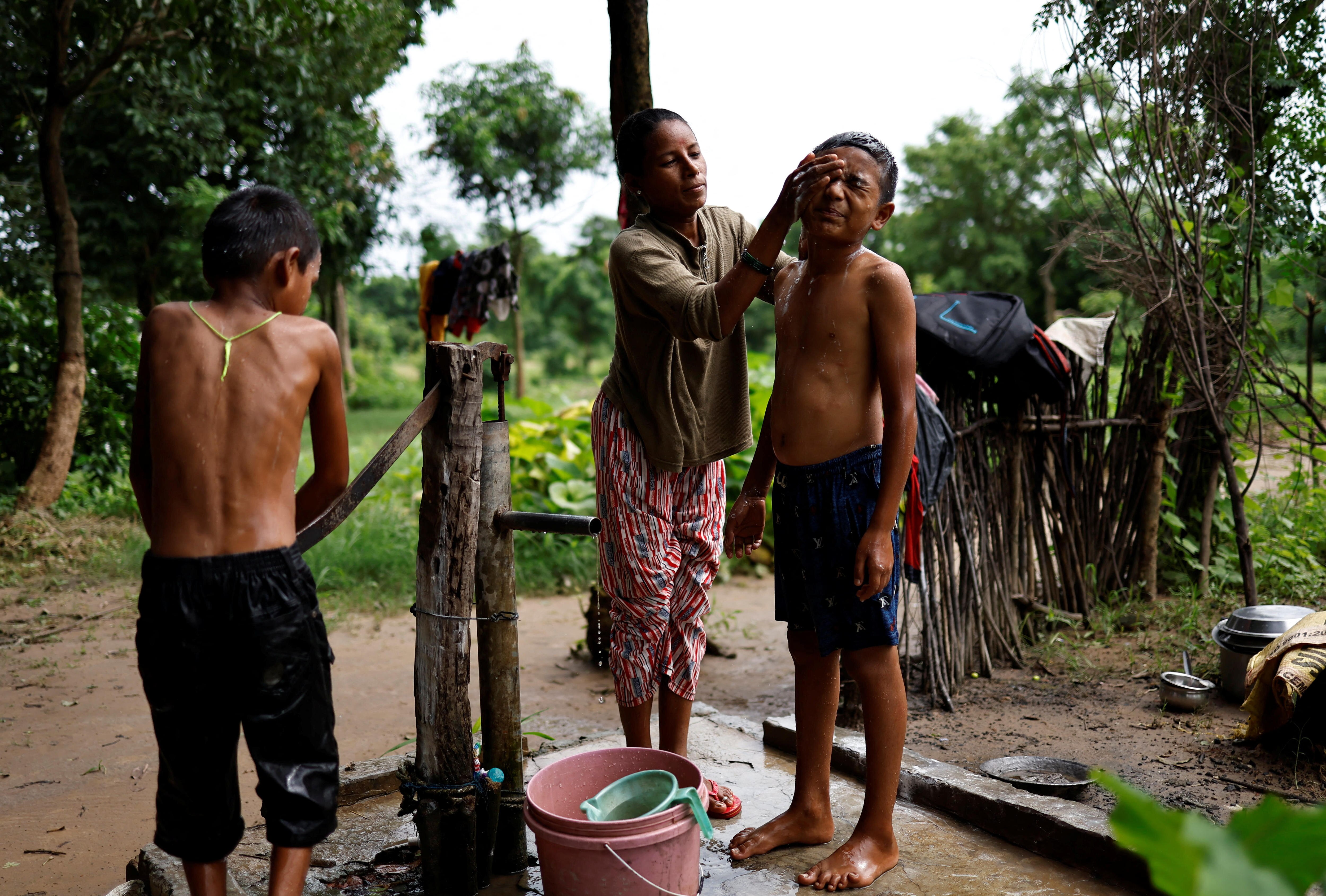 A woman washes a boys face with water while another boy works a hand pump. Trees, wooden fence and fields behind them