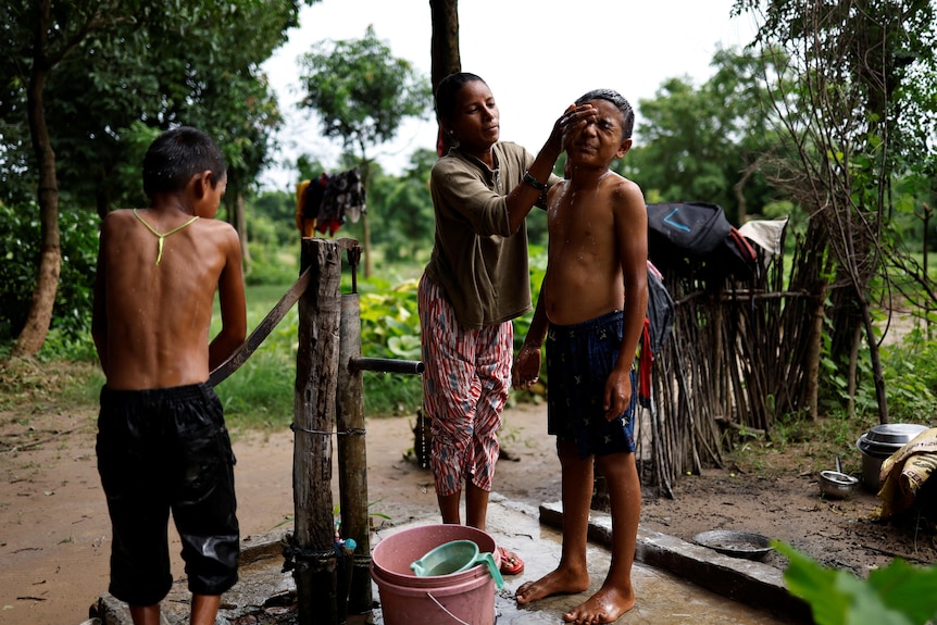 A woman washes a boys face with water while another boy works a hand pump. Trees, wooden fence and fields behind them