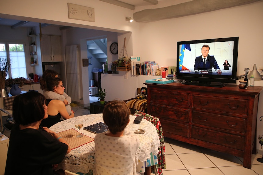 Una familia se sienta alrededor de una mesa redonda de comedor viendo la televisión mientras Macron habla.