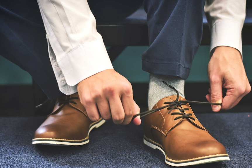 A man ties laces on a pair of brown leather shoes to depict a basic item from a men's workwear wardrobe.