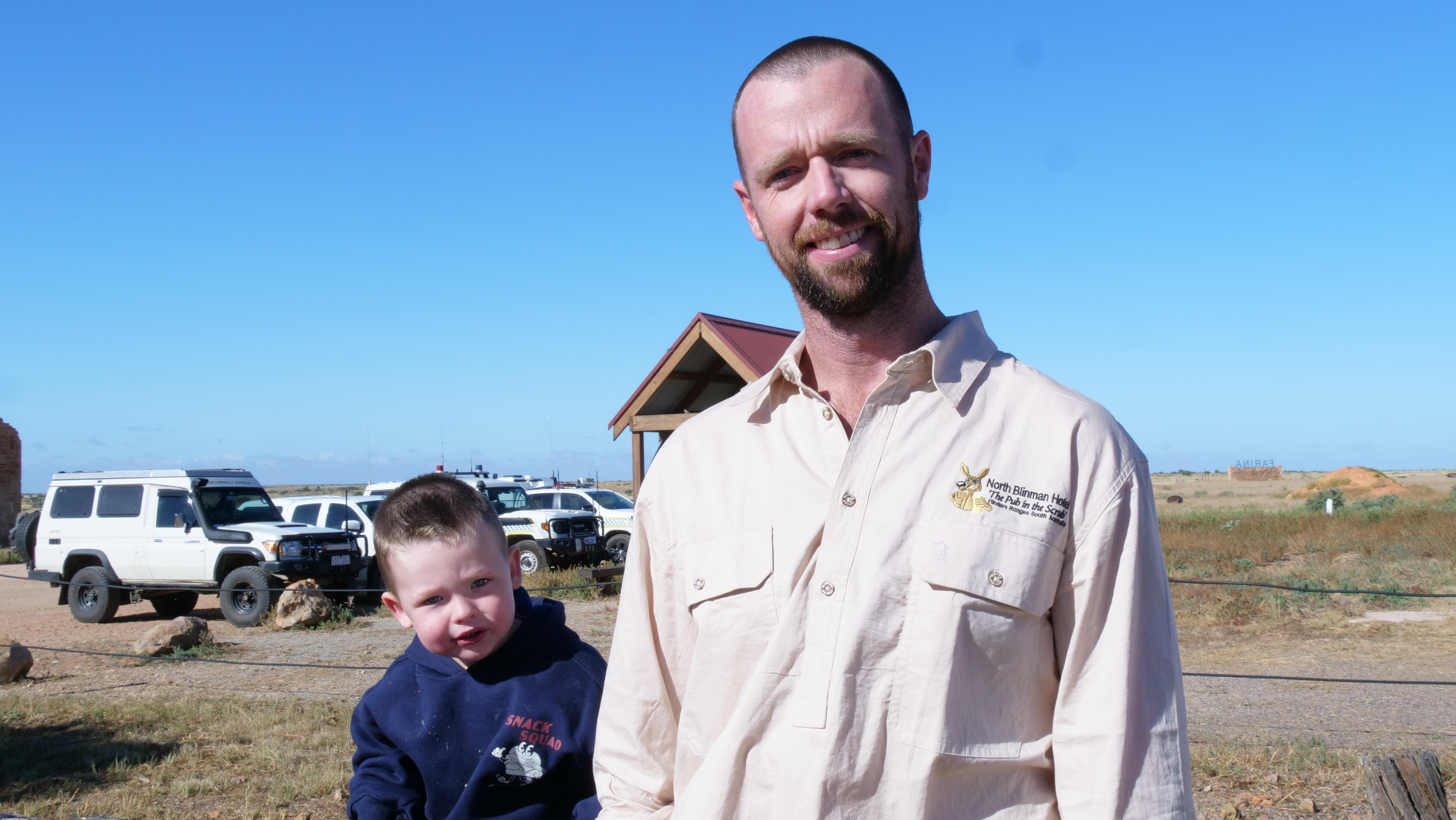 A young man and his small son smiling at the camera on a sunny day.