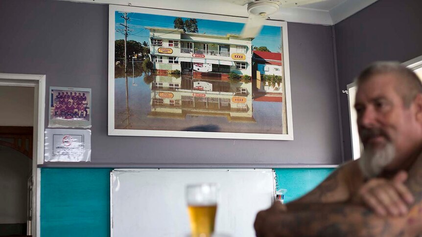 A man drinks at a bar, with a photo of the flooded-out hotel on the wall behind him.