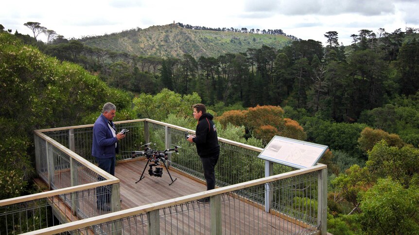 Two people standing on a wooden deck with forest in the background, with a small drone aircraft beside them.