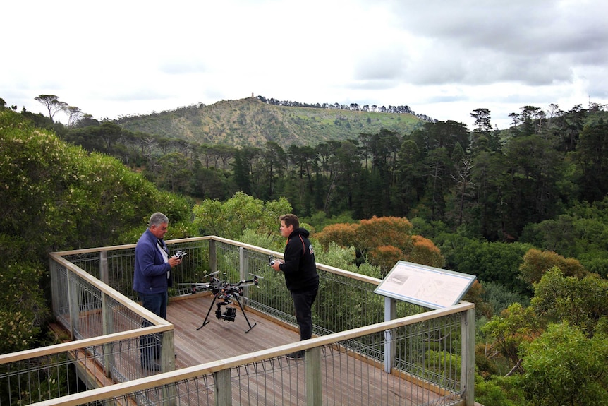 Two people standing on a wooden deck with forest in the background, with a small drone aircraft beside them.
