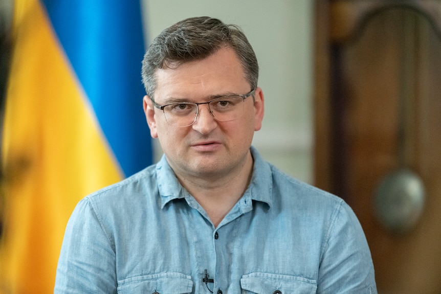 Close up of spectacled man in blue button up shirt sits in front of Ukrainian flag.