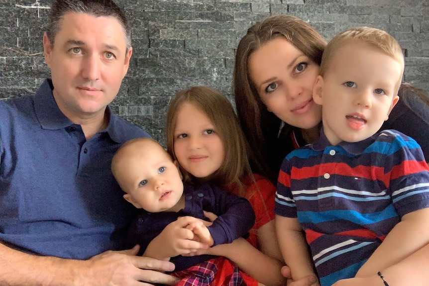 Owen Stuchbery, left, sits with his three children and his wife, far right, against a grey brick wall.