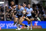 A Cronulla NRL player stumbles forward carrying the ball as he is tackled from behind by two defenders.