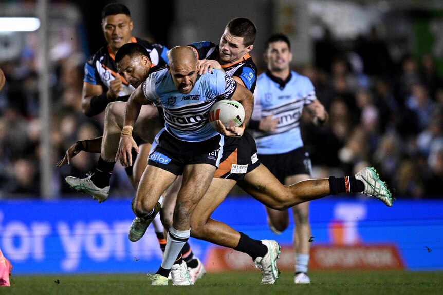 A Cronulla NRL player stumbles forward carrying the ball as he is tackled from behind by two defenders.