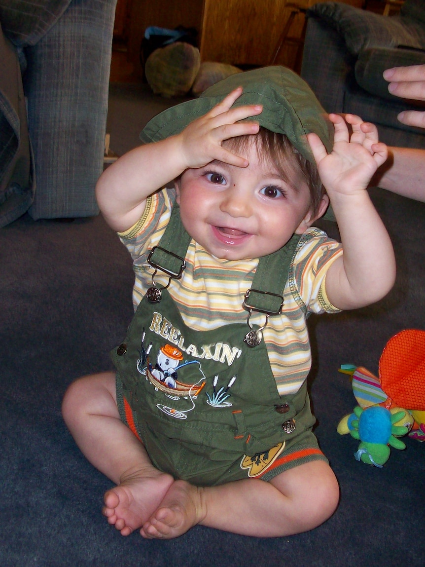 A baby with brown eyes in a green hat and overalls 