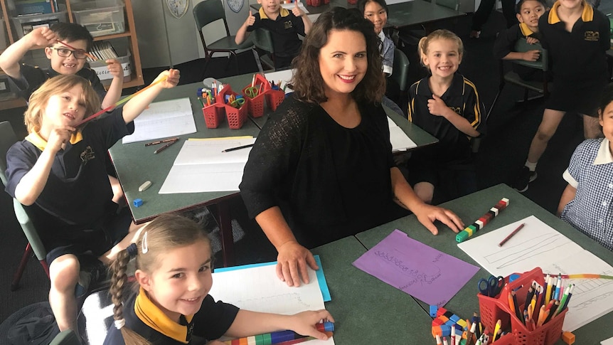 Graduate teacher Elise Andrew sits down behind a desk in her classroom surrounded by primary school students.