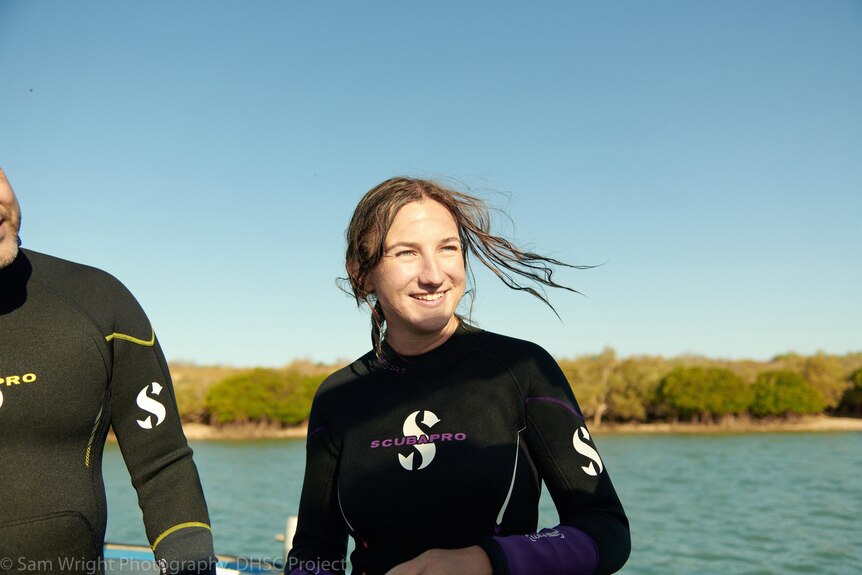 Young woman wearing a wetsuit stands on a boat looking to the right of the camera and smiling.
