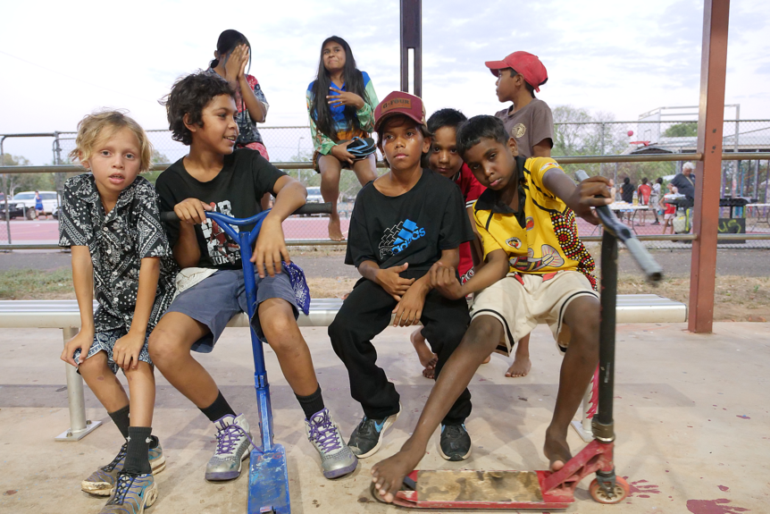A group of Indigenous children sit on spectator seats at a basketball court