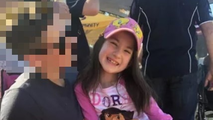 An eight-year-old girl with dark hair and wearing a pink cap sits on the knee of a person whose face is blurred.